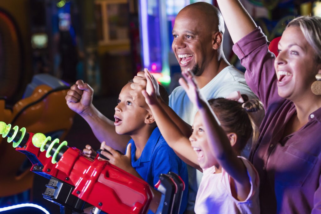 Family entertainment center franchise - An interracial family having fun together playing at an amusement arcade. The African-American father and Caucasian mother are in their 30s. The 9 year old boy and 5 year old girl are sitting in their parents' laps playing a video arcade game. The main focus is on father and son. Everyone is cheering after winning the game.