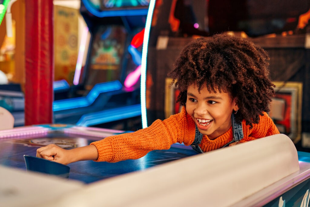 FEC franchise support - A cute African-American child with afro curls playing air hockey at an amusement park and carousel on her day off in the evening.