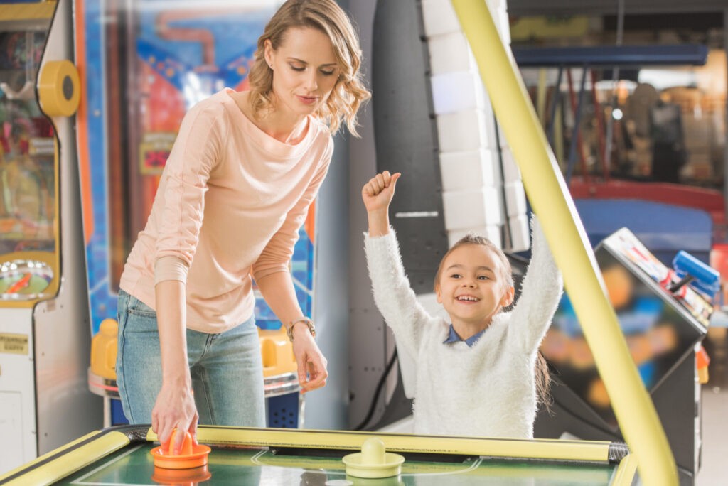 smiling mother and triumphing little daughter playing air hockey in family entertainment center